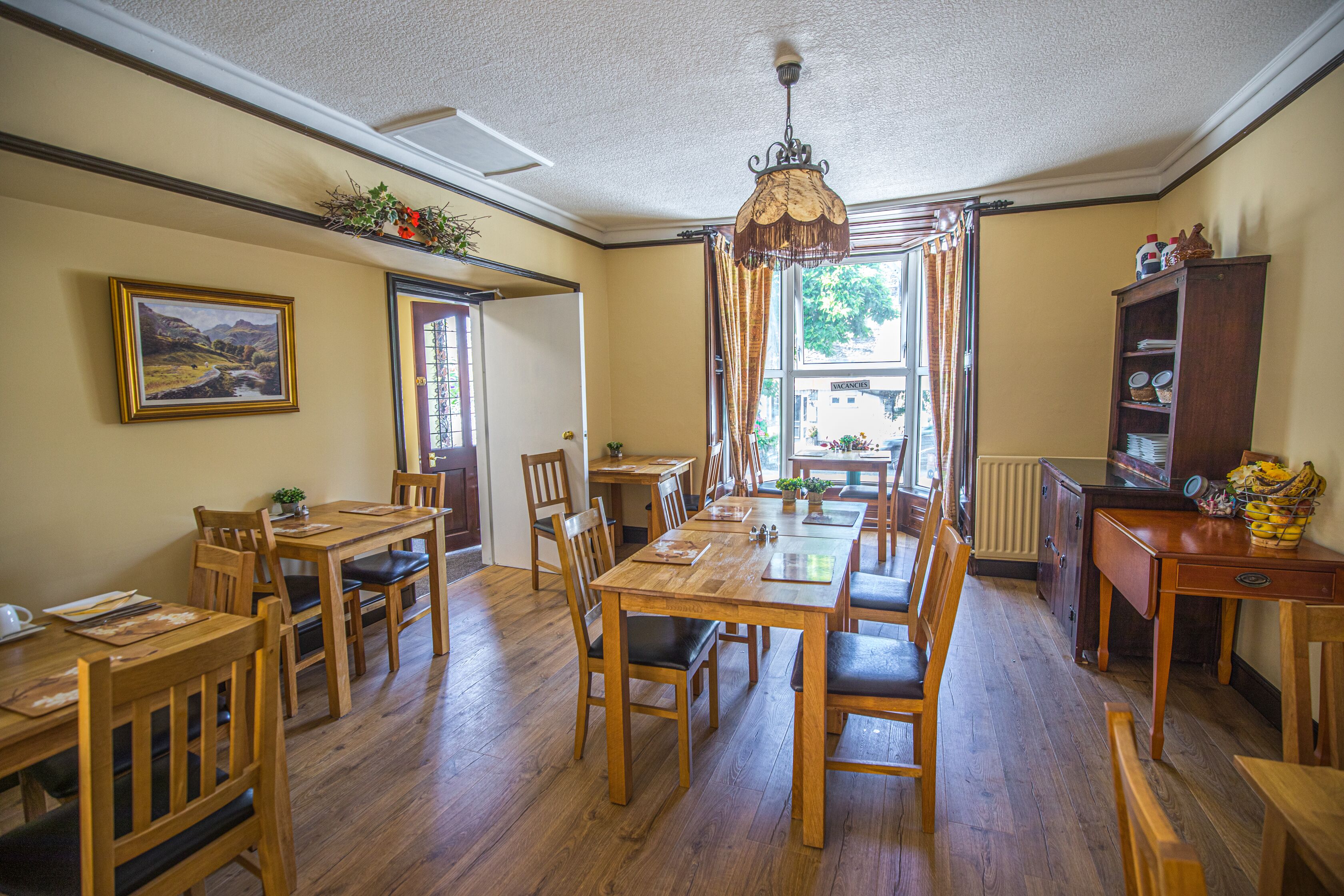 A dining area with wooden tables and chairs, a chandelier, and a view of the outdoors through large windows.