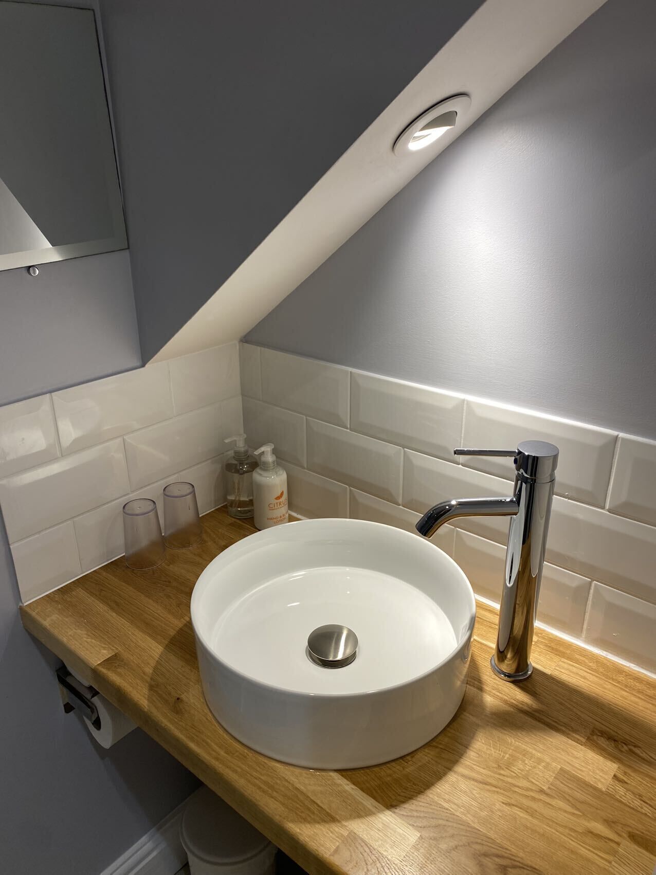 A modern bathroom with a round white sink, chrome faucet, and wooden countertop. There are two bottles of hand soap and a roll of toilet paper on the counter.