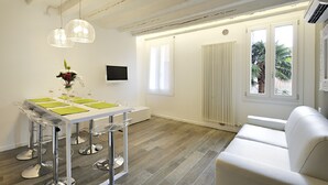 Un comedor moderno con una mesa blanca, cuatro taburetes de bar y un sofá blanco. Hay un televisor en la pared y una ventana con vista a palmeras.
