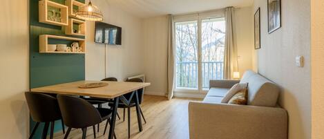 A modern living room with a wooden dining table, black chairs, a beige sofa, and a large window with a view of trees.
