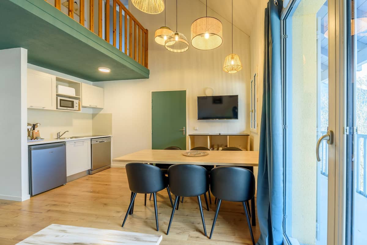 A modern kitchen with white cabinets, a microwave, and a dishwasher. There is a dining area with a wooden table and black chairs. A television is mounted on the wall above the table.