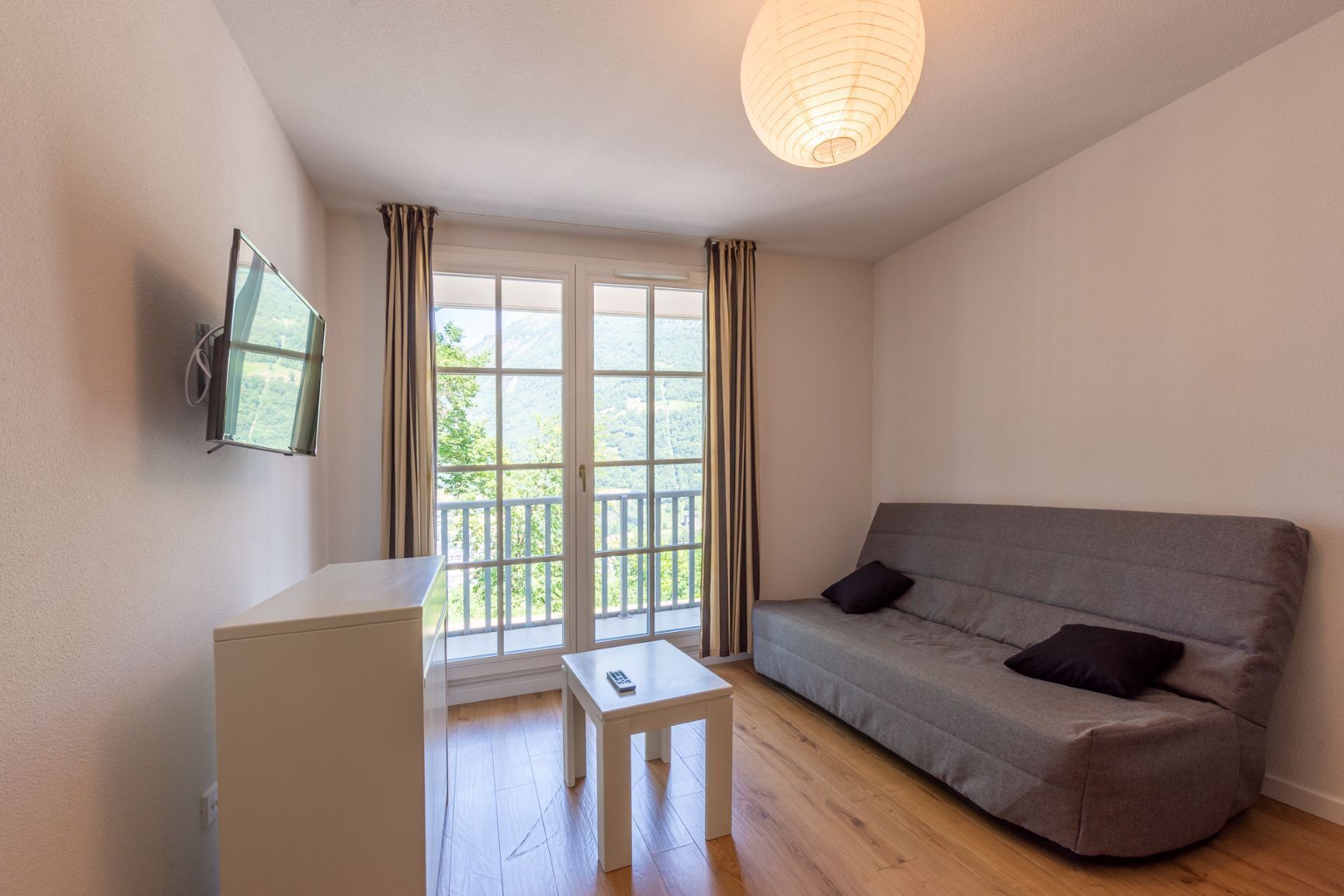 Une pièce avec un canapé gris, une petite table blanche, un téléviseur fixé au mur et un balcon avec des rideaux.