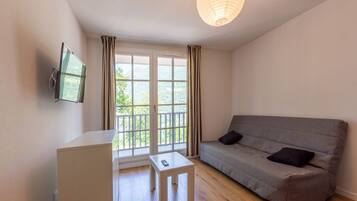 Une pièce avec un canapé gris, une petite table blanche, un téléviseur fixé au mur et un balcon avec des rideaux.