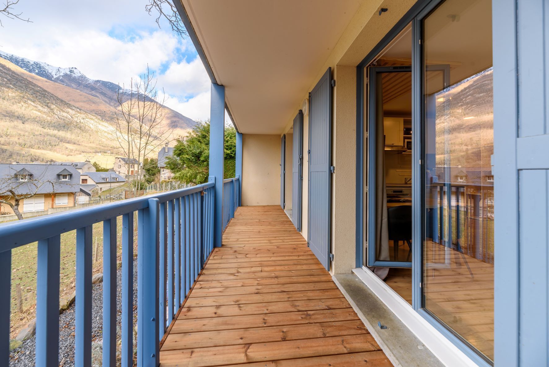 Una terraza de madera con barandilla azul, una puerta de cristal que da a una habitación y vistas a un paisaje montañoso.