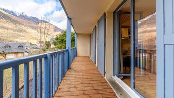 Una terraza de madera con barandilla azul, una puerta de cristal que da a una habitación y vistas a un paisaje montañoso.