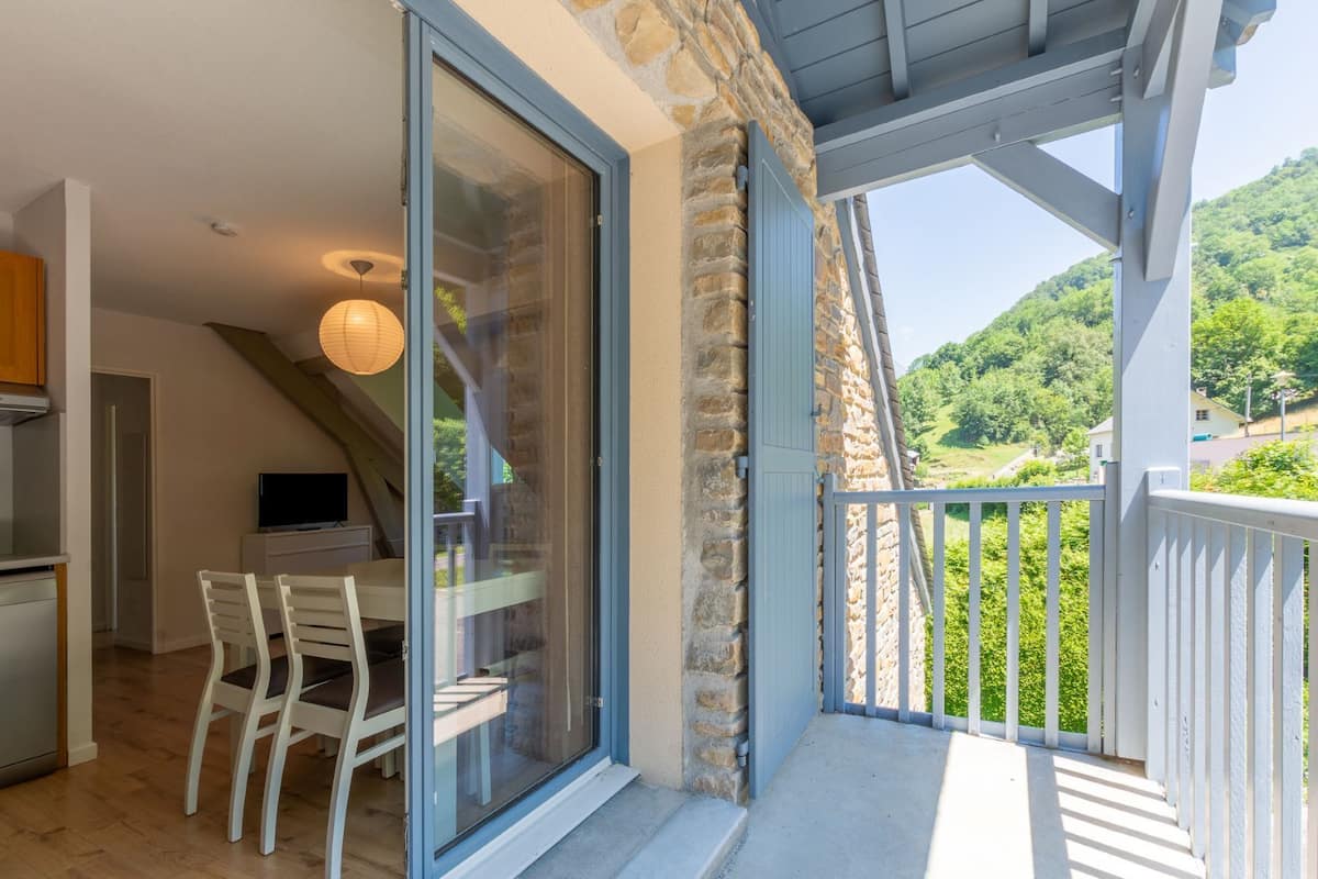 A balcony with a stone wall, a dining area with a table and chairs, and a view of a green landscape.