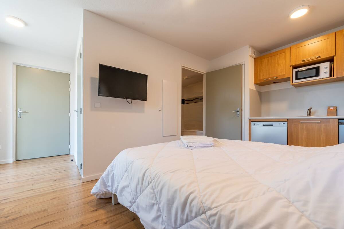 A bedroom with a large bed, a flat-screen TV mounted on the wall, and a kitchenette with a microwave and a dishwasher.