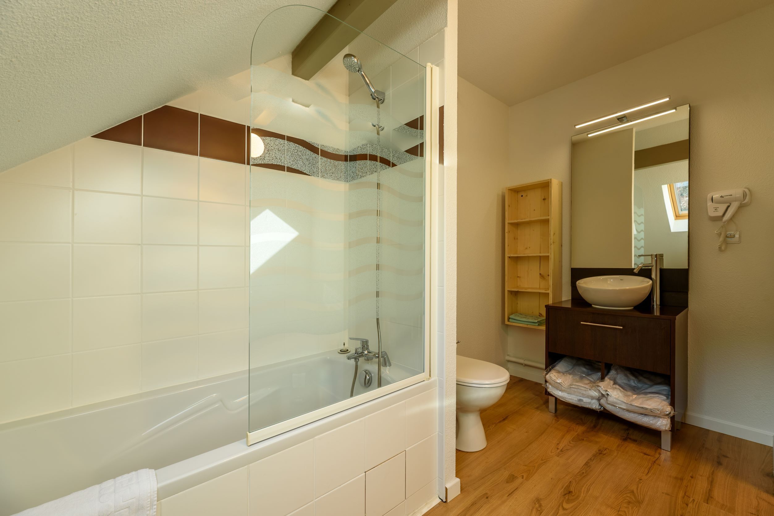 Un baño moderno con ducha envuelta en vidrio, inodoro blanco y tocador de madera con espejo.