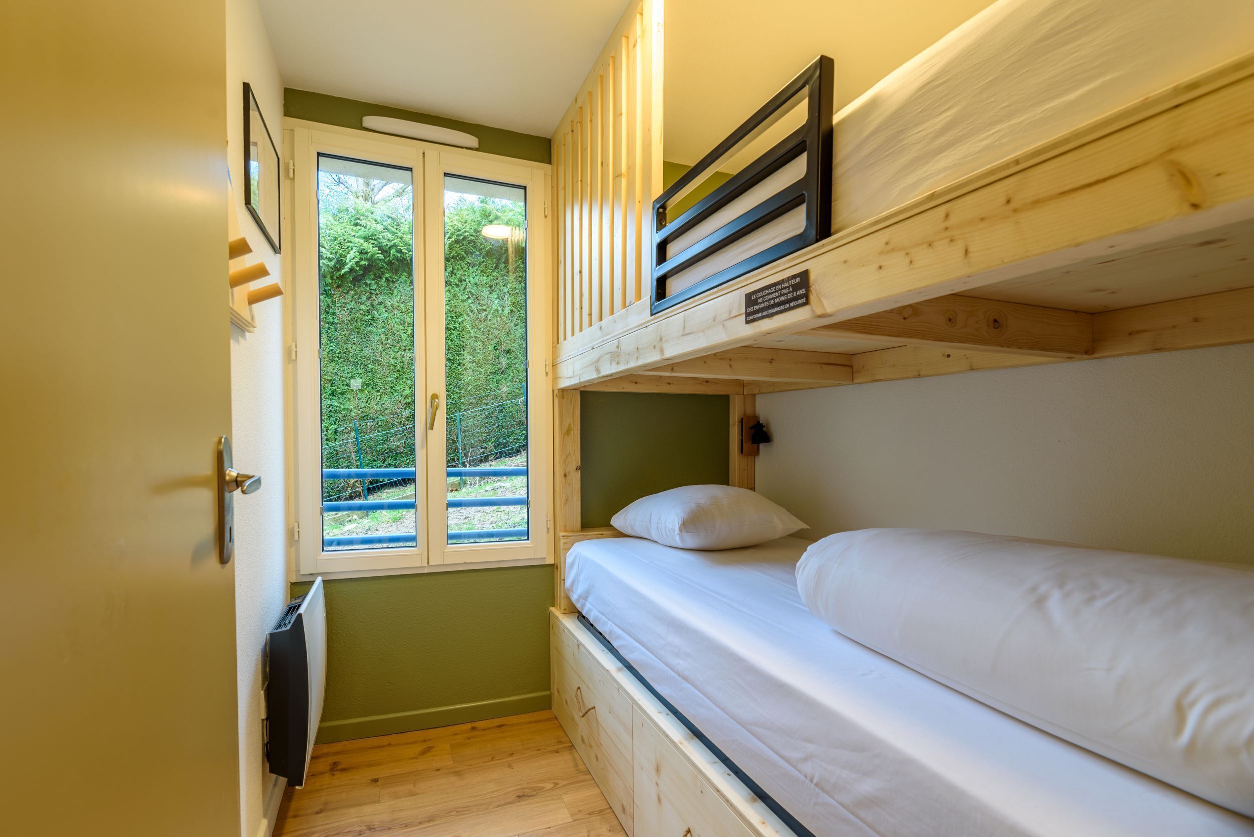 Una habitación pequeña con literas, una ventana que da a zonas verdes y una puerta con manija.