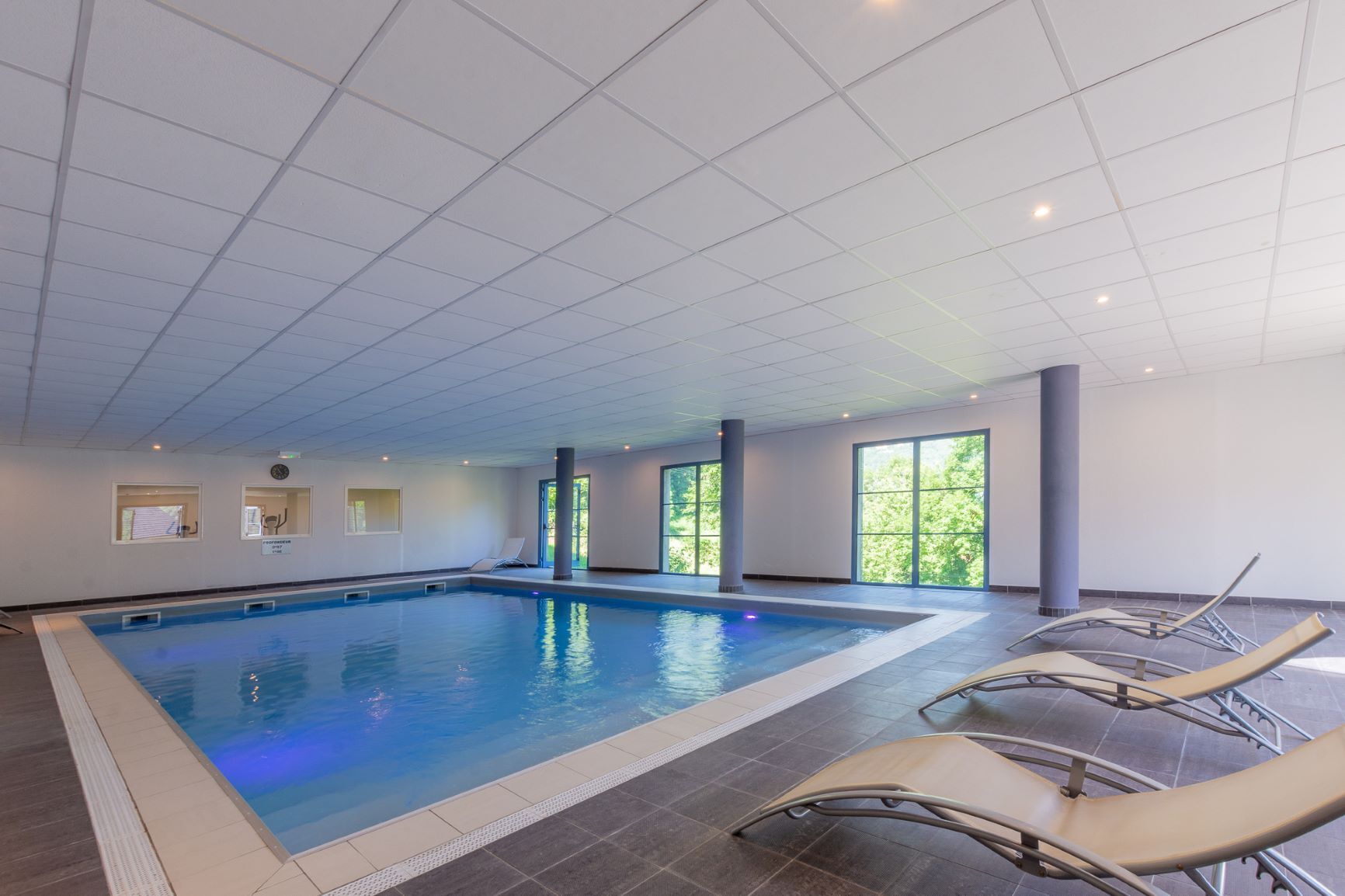 Une piscine intérieure avec des parois en verre transparent, un sol carrelé et quelques chaises longues.