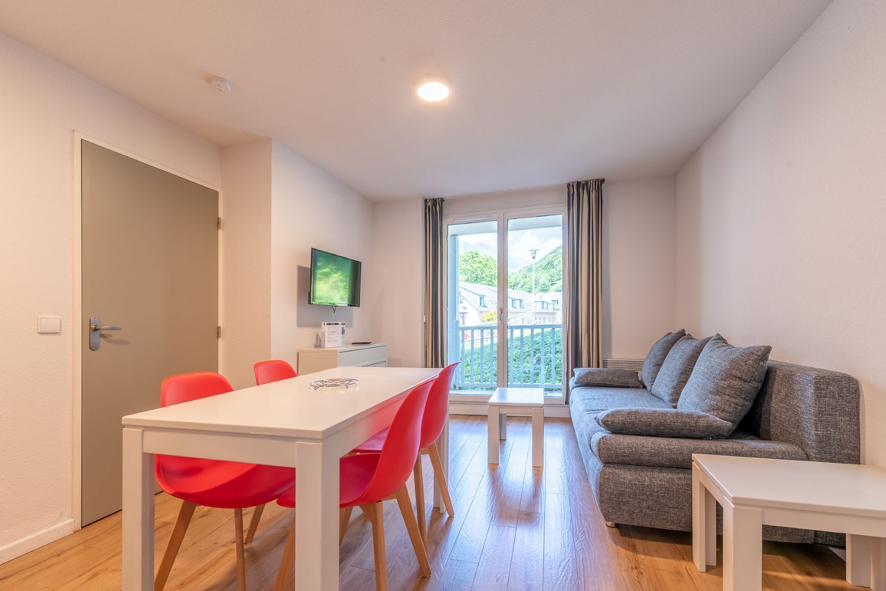 A modern living room with a white table and red chairs, a grey sofa, and a TV on the wall.