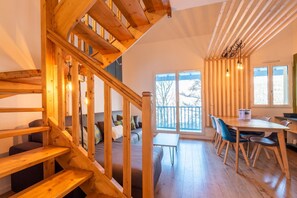 A wooden staircase leading to a balcony with a view, a dining area with chairs and a table, and a cozy seating area with cushions.