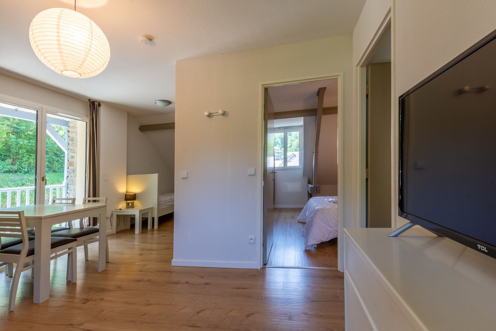 Ein Zimmer mit Holzboden, einem Bett, einem Schreibtisch mit Stuhl, einem Tisch mit Stühlen, einer Pendelleuchte und einem Fernseher.