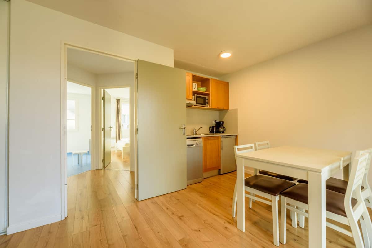 A compact kitchen with wooden cabinets, a microwave, and a coffee maker. There is a dining area with a white table and chairs.