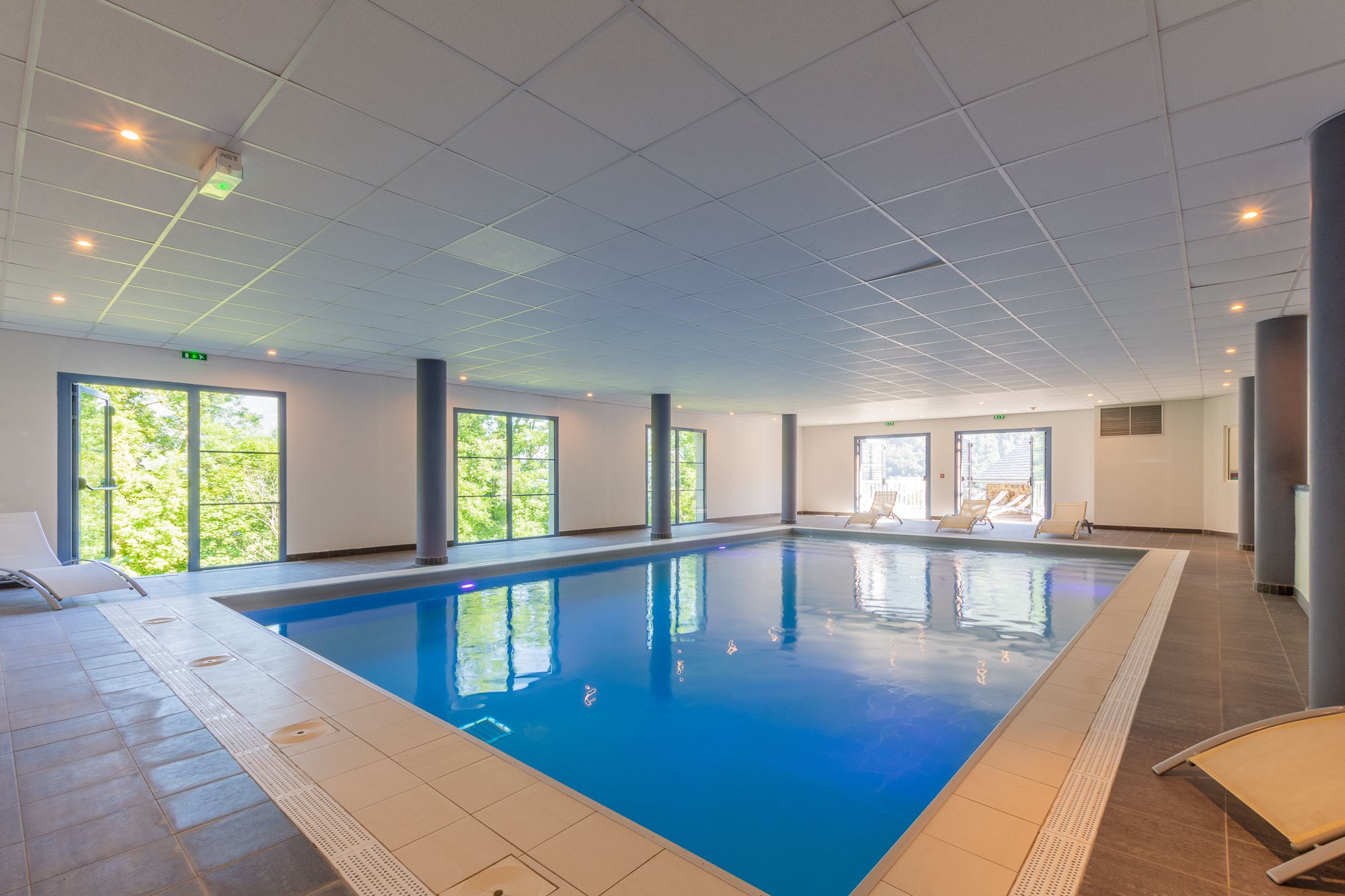 An indoor swimming pool with clear glass walls, a tiled floor, and a ceiling with recessed lighting.