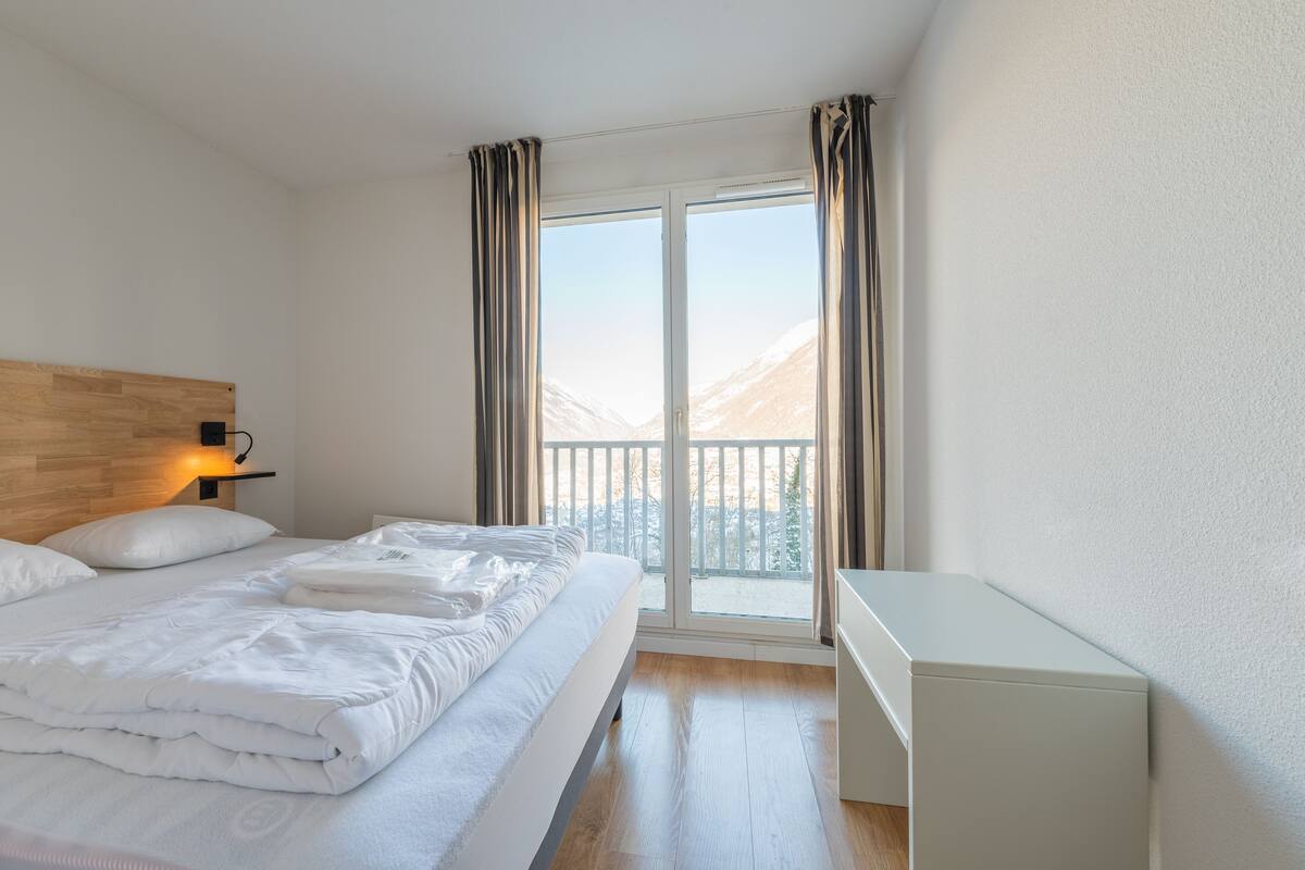 A bedroom with a bed, a nightstand, a desk, and a window with curtains.