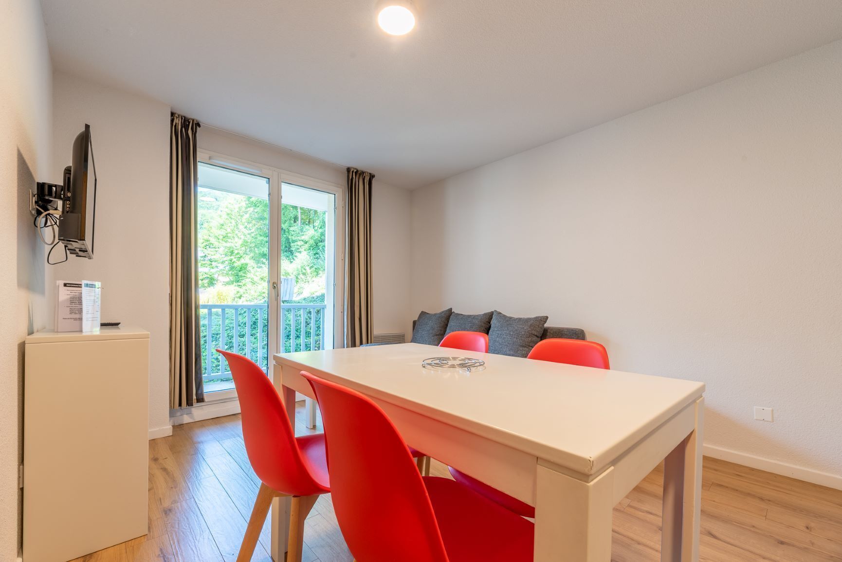 Una habitación moderna con una mesa blanca, sillas rojas, un sofá con cojines grises y un ventanal corredero que da a un balcón.