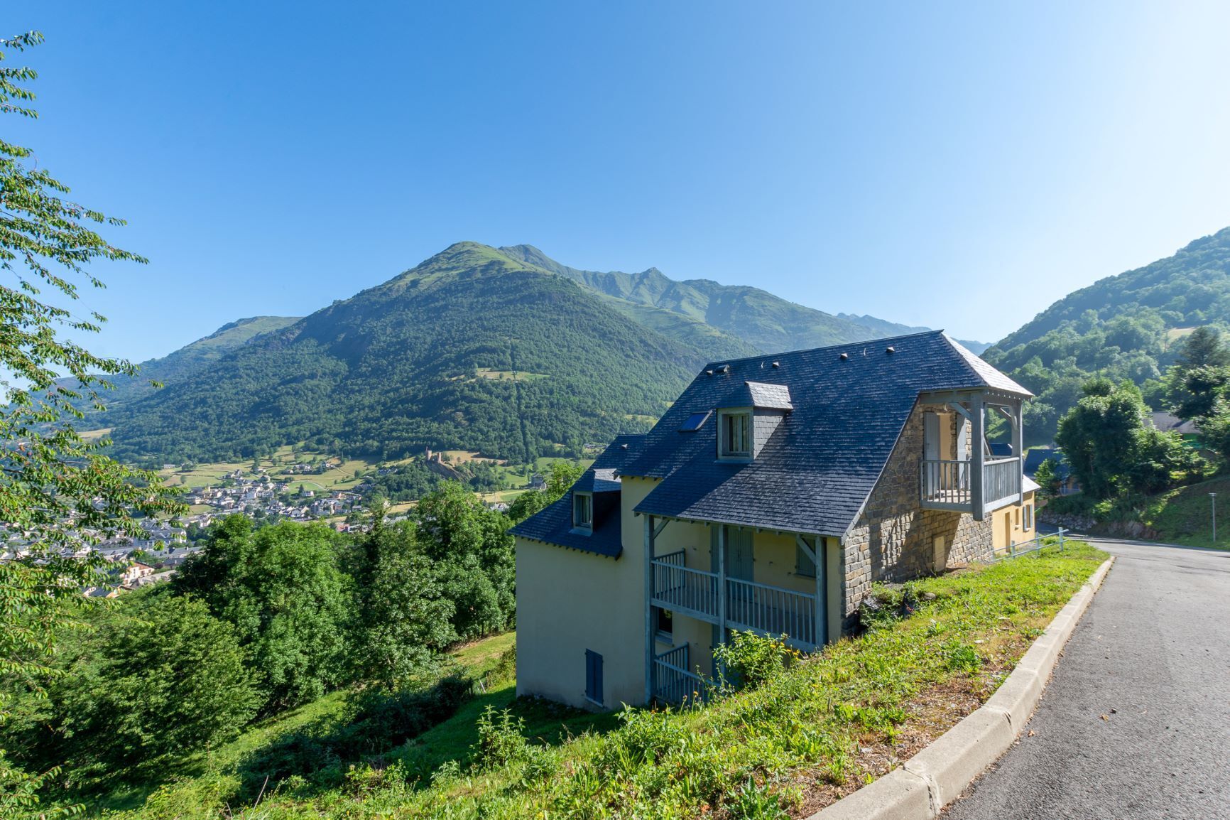 Une maison dotée d’un balcon, entourée de verdure et avec une montagne en toile de fond.