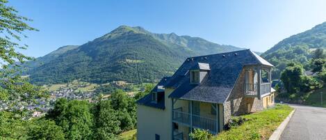 Une maison dotée d’un balcon, entourée de verdure et avec une montagne en toile de fond.