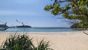 Una playa con agua turquesa cristalina, orilla arenosa y algunos barcos a lo lejos.