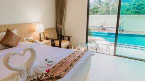 Habitación de hotel con cama, escritorio, silla, mesa de vidrio y vistas a una piscina y follaje exterior.