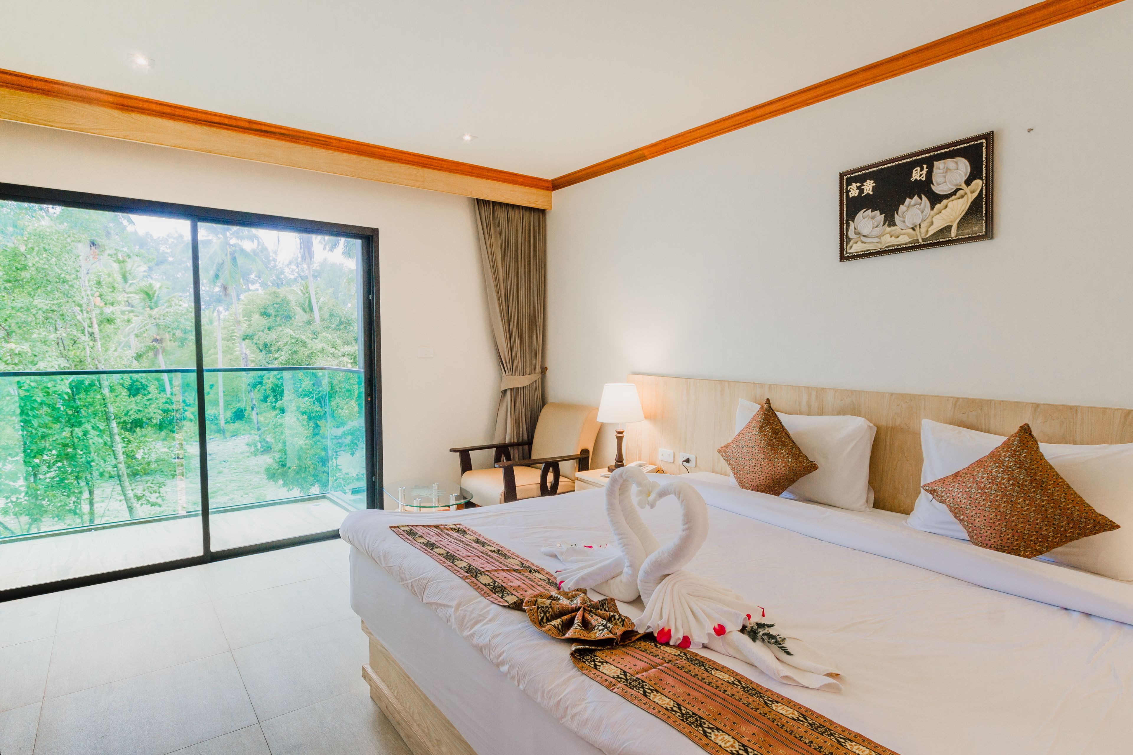 A hotel room with a large bed, a wooden headboard, a nightstand, a lamp, and a view of greenery through a sliding glass door.