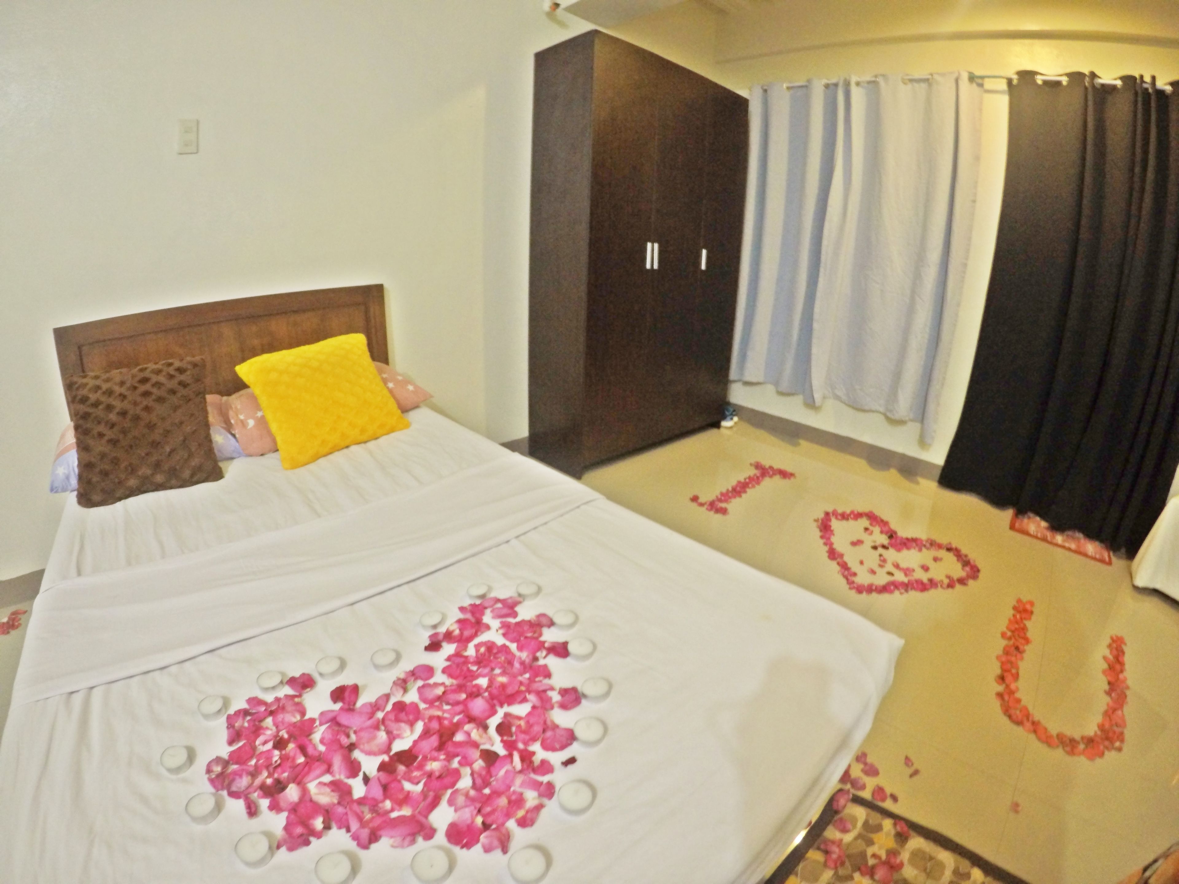 A bed with a white bedspread, a yellow pillow, and pink rose petals arranged on the bed.