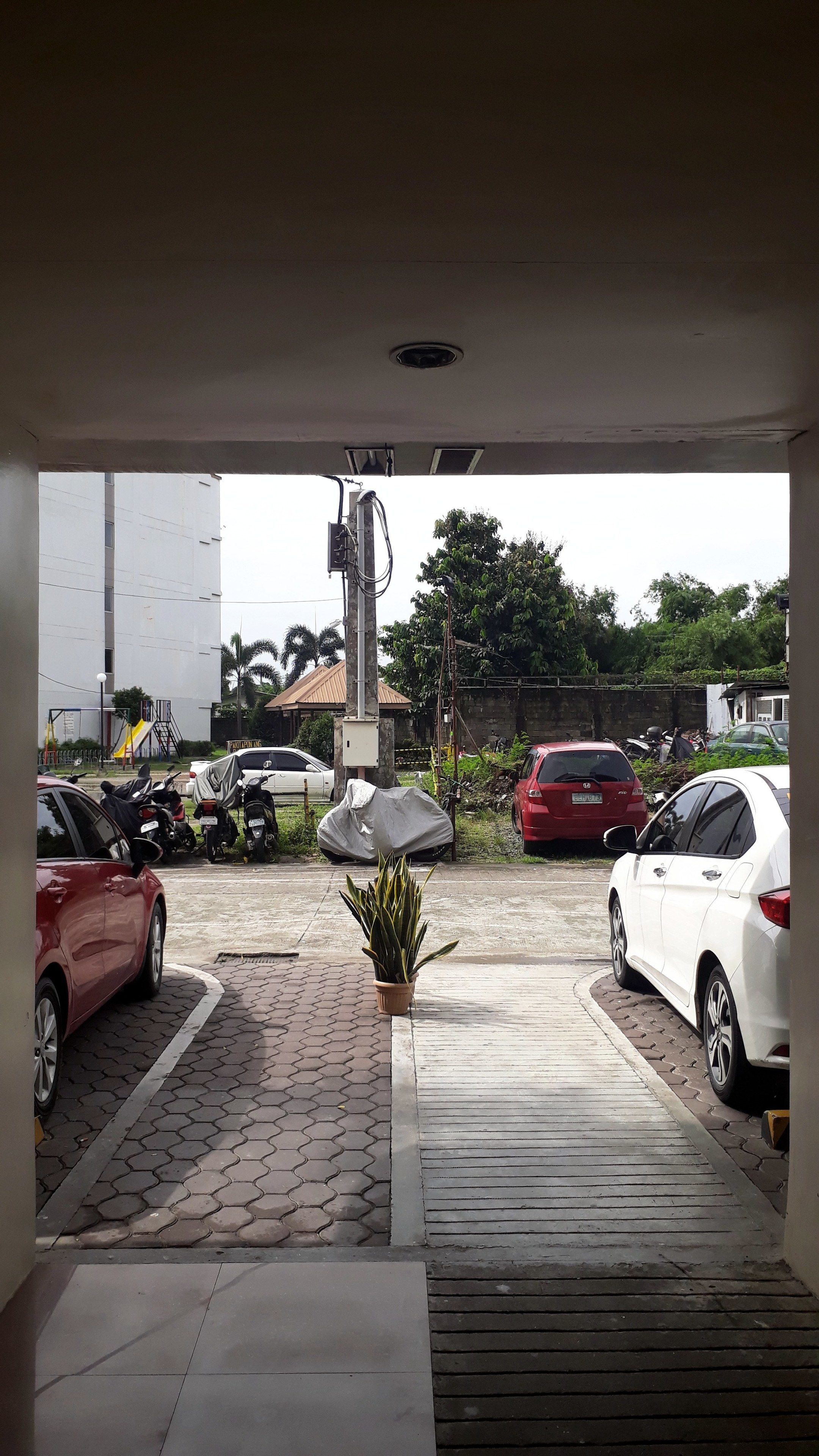 Un área de estacionamiento con varios coches y motocicletas, una zona techada y una planta en maceta.