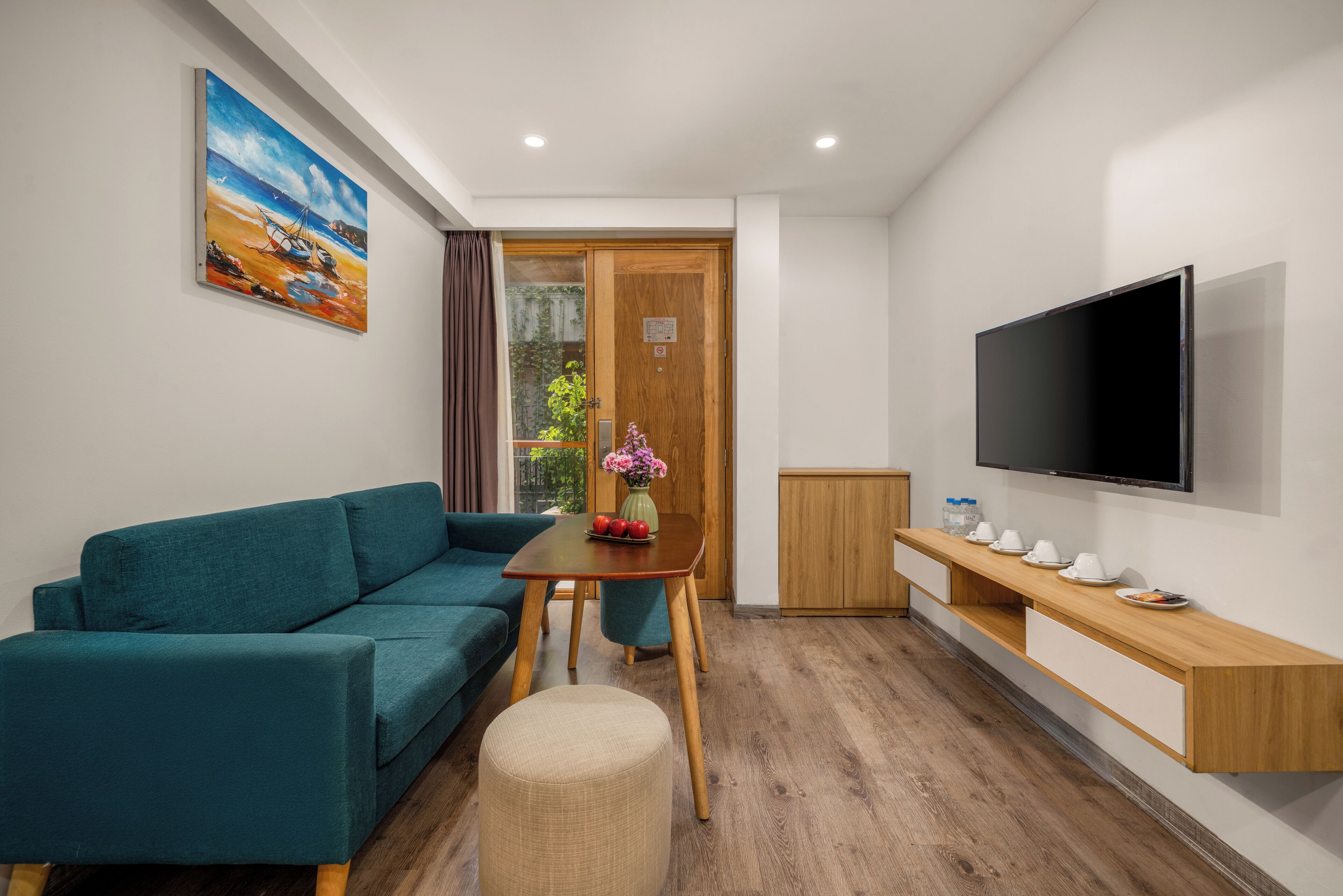 Ein modernes Zimmer mit einem türkisfarbenen Sofa, einem Holztisch mit einer Vase und Obst, einem an der Wand montierten Fernseher und einem Holzkasten.