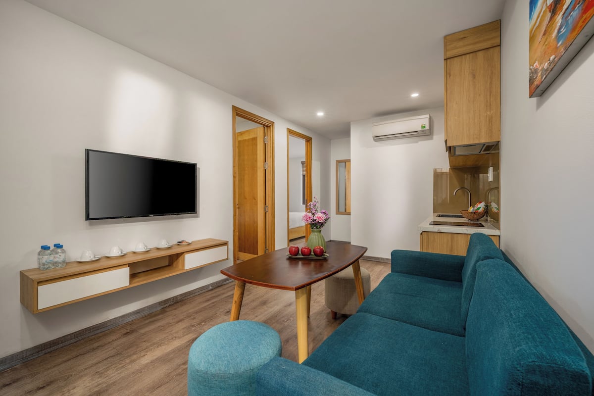 A modern living room with a flat-screen TV, wooden furniture, a teal sofa, and a small table with decorative items.