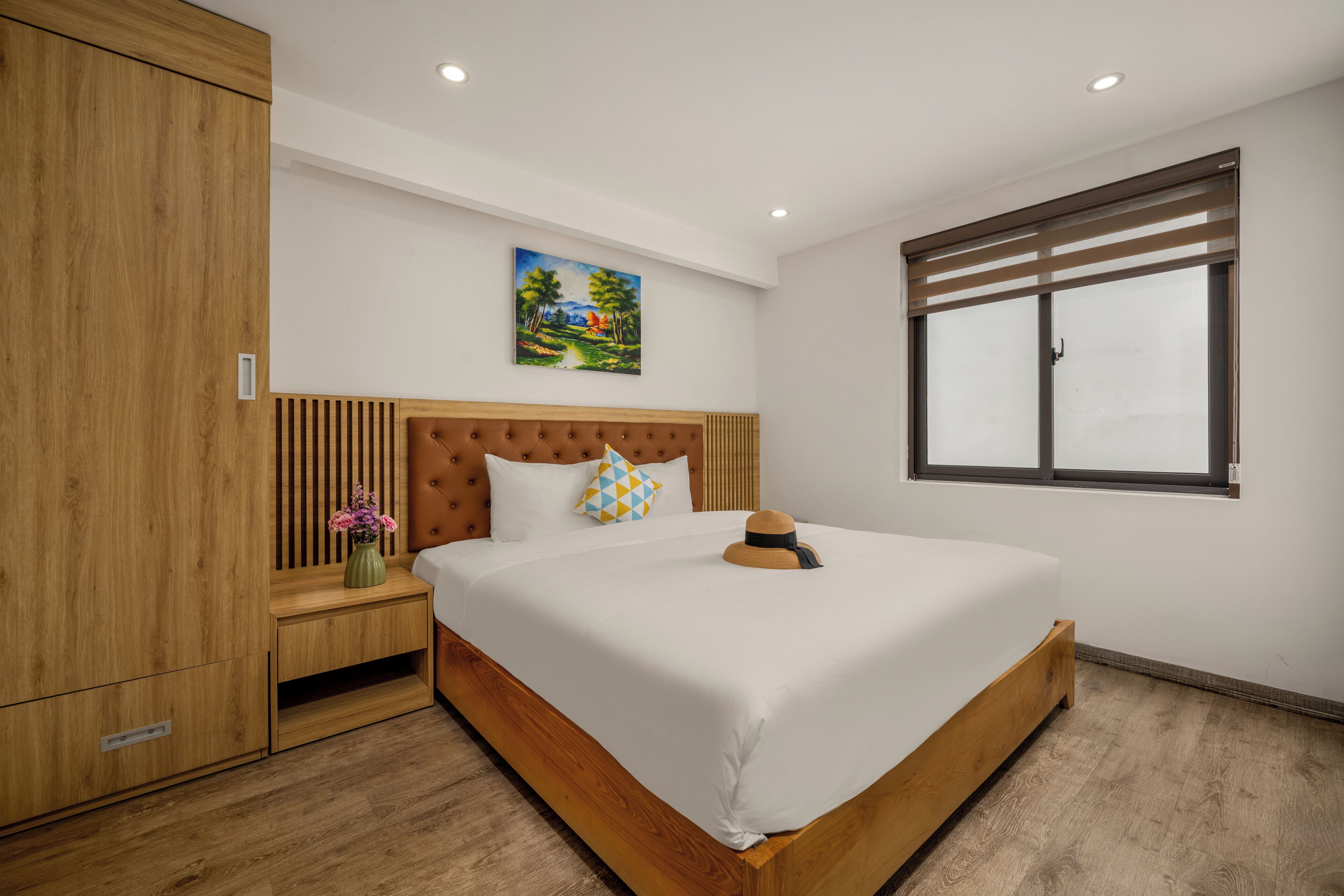 Een moderne slaapkamer met een houten bed, een groot raam met zonwering, een nachtkastje met een bloevas, en een schilderij aan de muur.