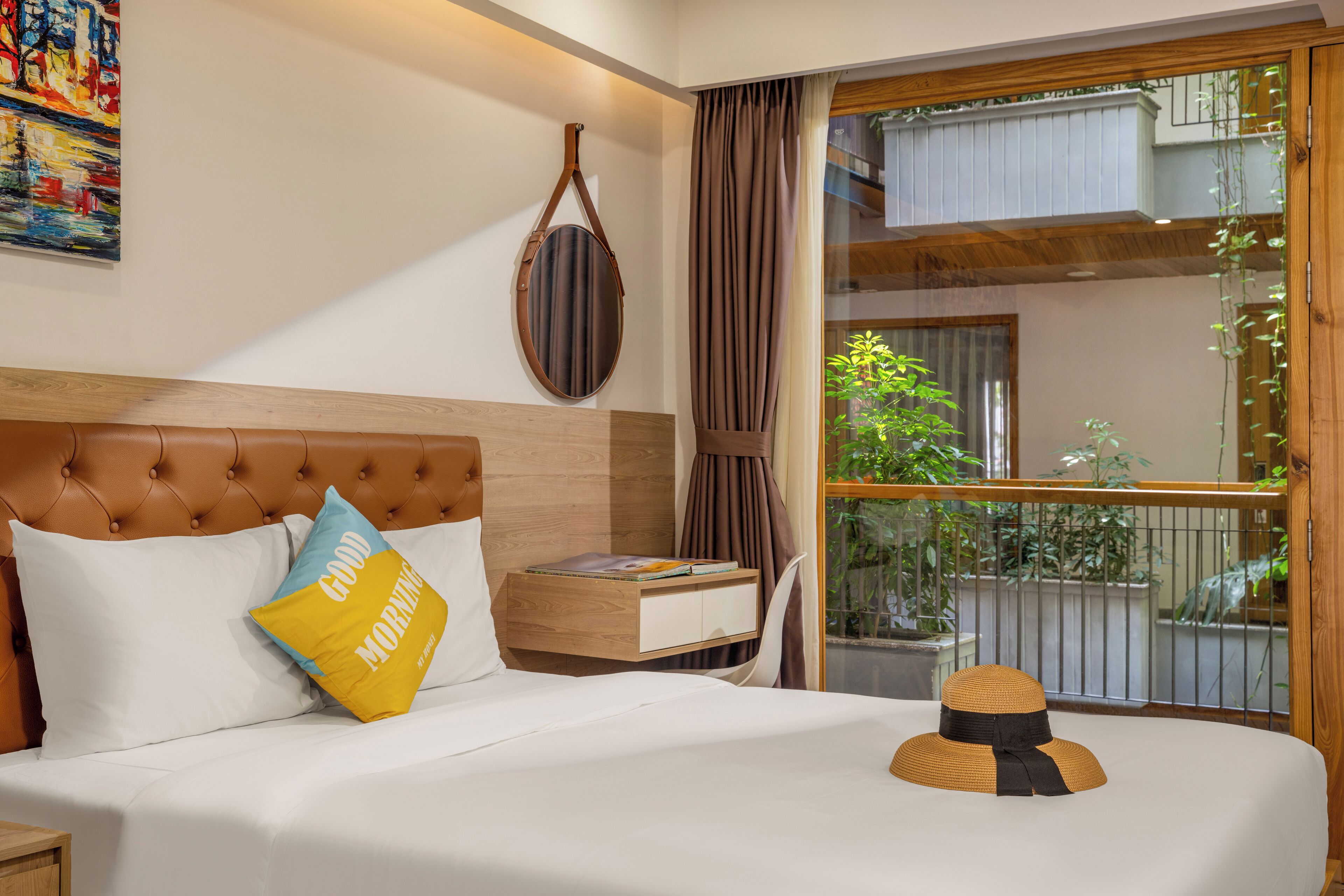 A neatly made bed with a pillow, a bedside table with a book, and a view of a balcony with plants.