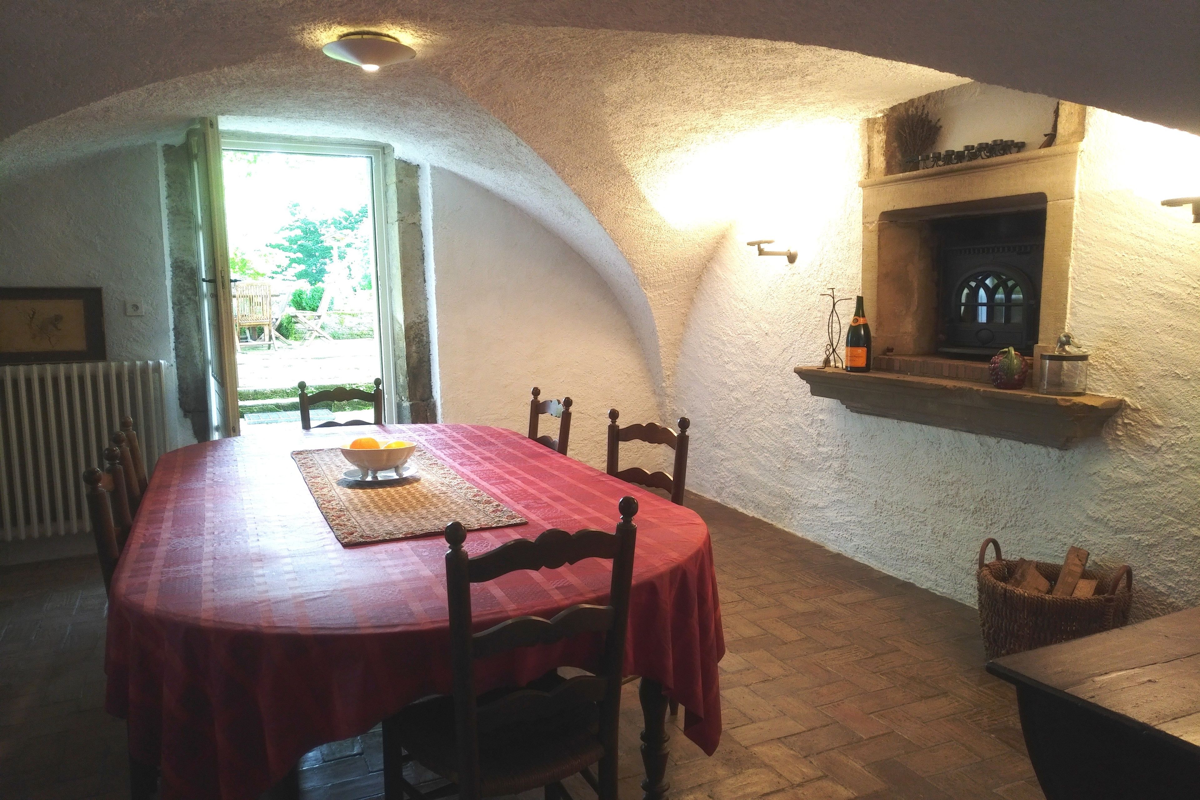A dining area with a red tablecloth, wooden chairs, a fireplace, and a basket of firewood.