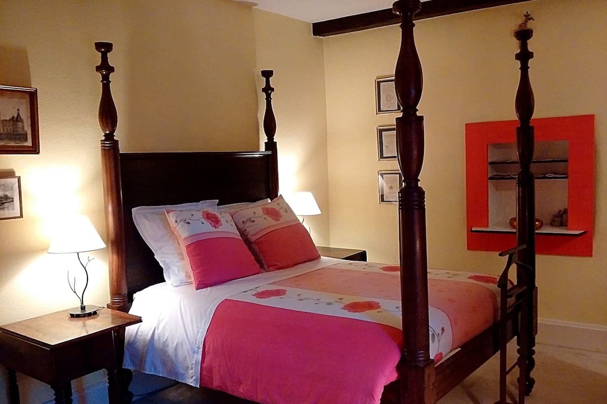Uma cama de dossel com colcha florida e almofadas cor-de-rosa, uma mesa de cabeceira com abajur e um espelho com moldura vermelha na parede.