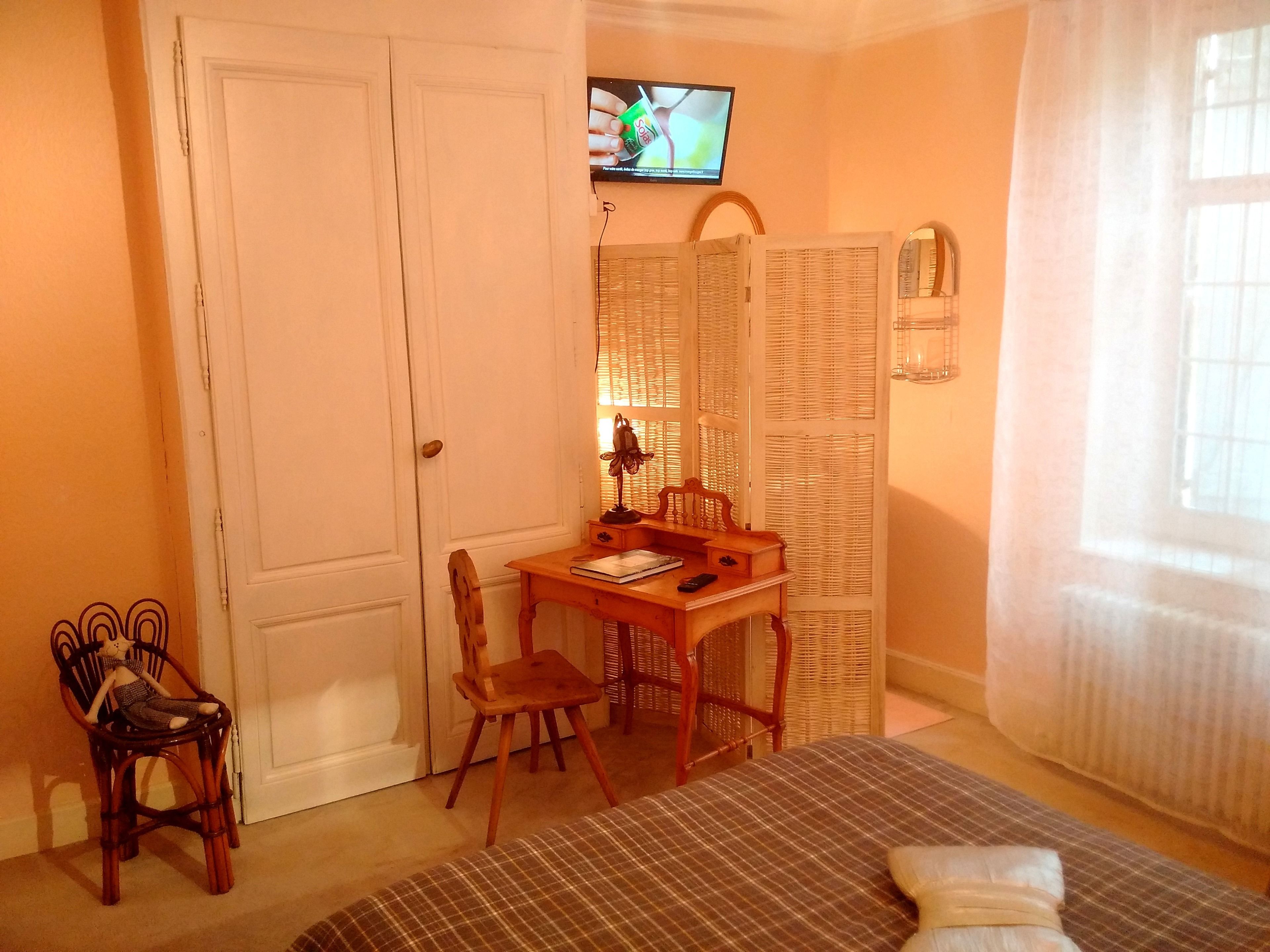 A room with a wooden desk, two chairs, a television on the wall, a wicker screen, and a window with curtains.
