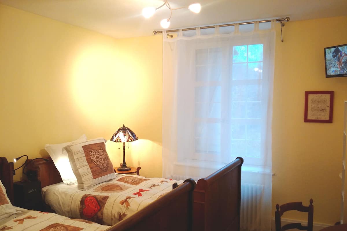 Quarto com duas camas, uma mesa de cabeceira com abajur, uma cadeira, uma janela com cortinas leves e um certificado emoldurado na parede.
