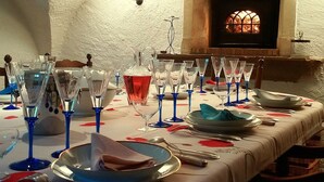 Una mesa de comedor dispuesta con vasos azules, platos y una jarrón con líquido rojo, con una chimenea al fondo.