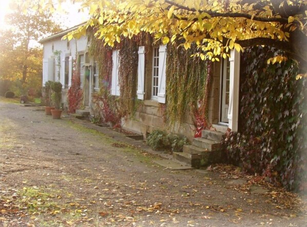 A building with a stone facade, white window frames, and a covered porch with yellow leaves. - Chambres d'hôtes Le Prieuré (Aydoilles)