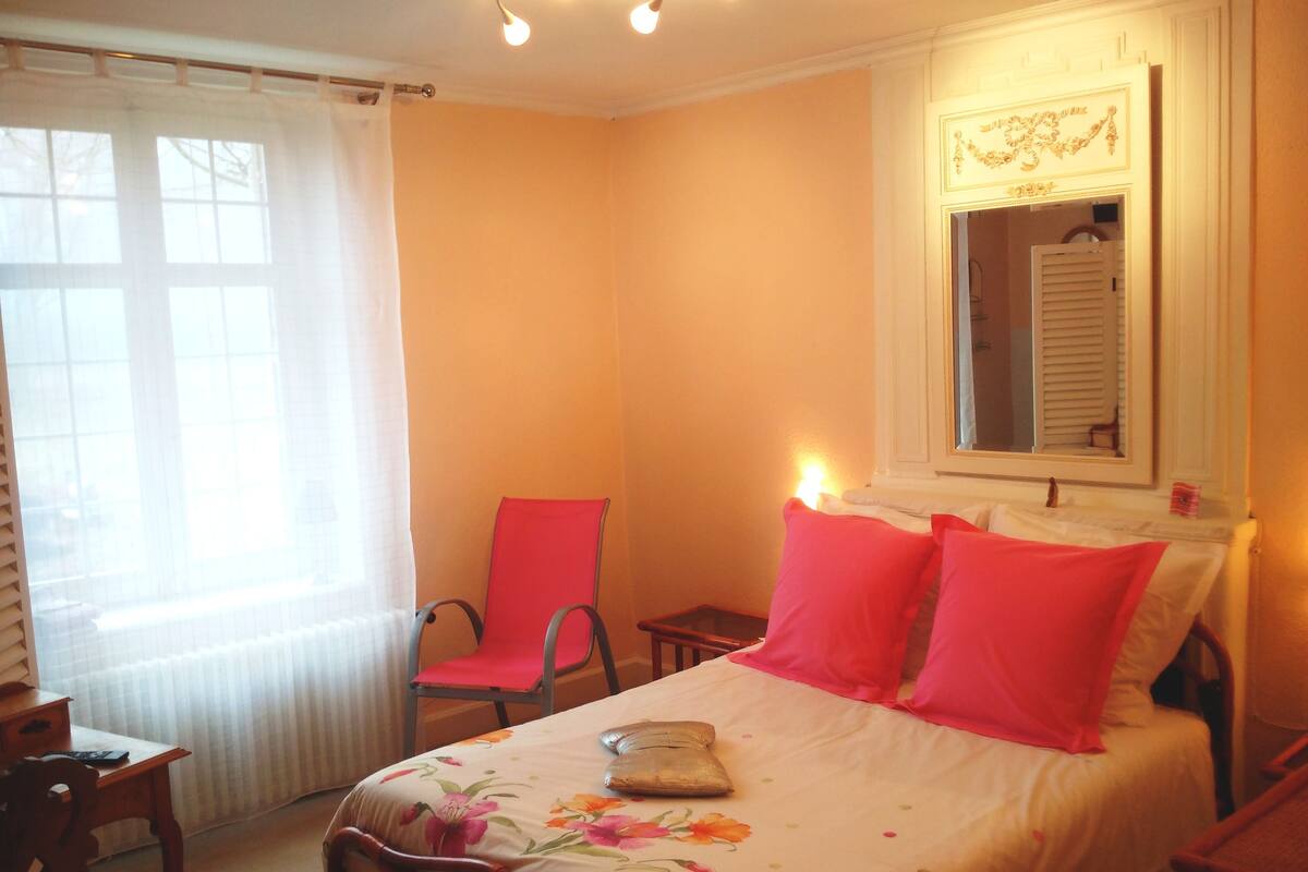 Quarto com cama, uma cadeira, uma mesinha e janela com cortinas.