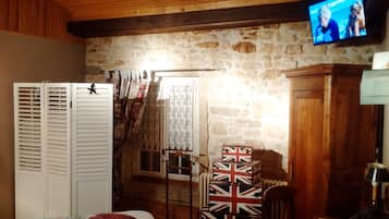 A room with a stone wall, wooden ceiling, a bed with a floral bedspread, a white wardrobe, a television mounted on the wall, and a stack of British flag-patterned boxes.