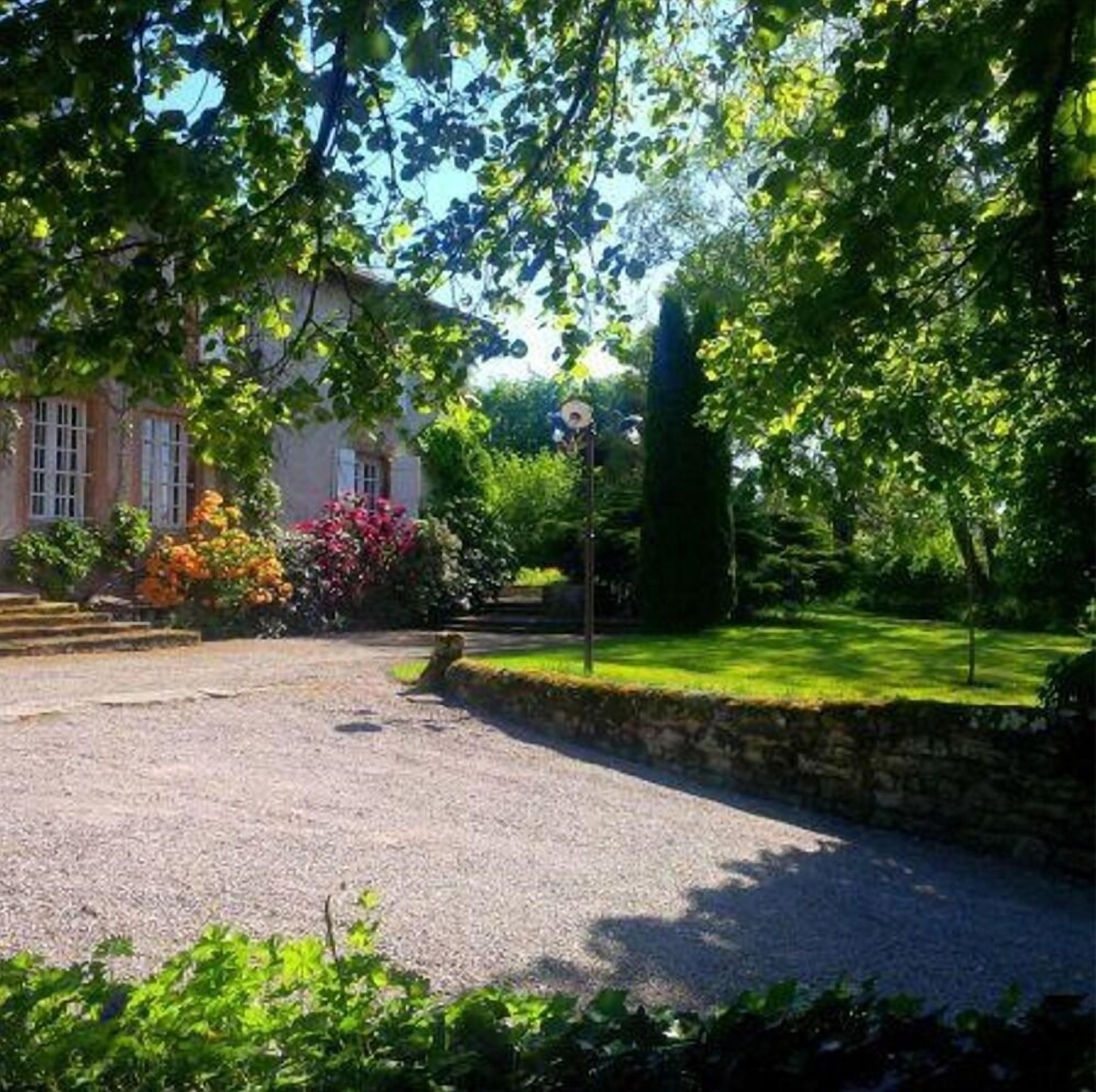 Une maison avec une allée en gravier, entourée d’une végétation luxuriante et de plantes en fleurs.