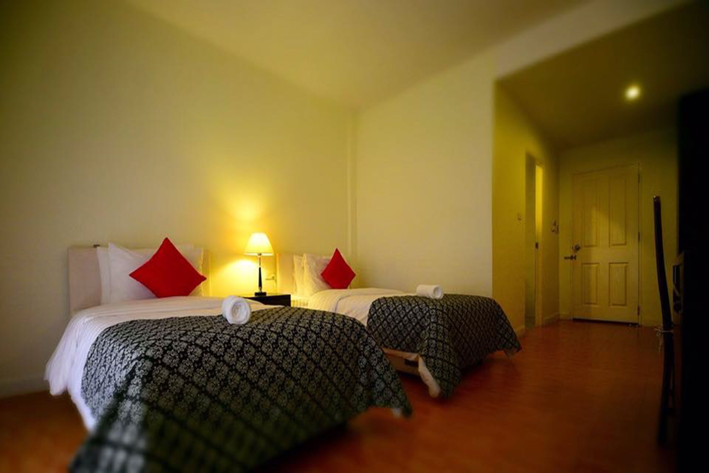 Quarto de hotel com duas camas, cada uma com roupa de cama estampada e almofadas vermelhas. Há uma mesa de cabeceira com uma luminária entre as camas.