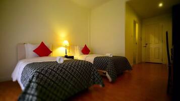 Quarto de hotel com duas camas, cada uma com roupa de cama estampada e almofadas vermelhas. Há uma mesa de cabeceira com uma luminária entre as camas.