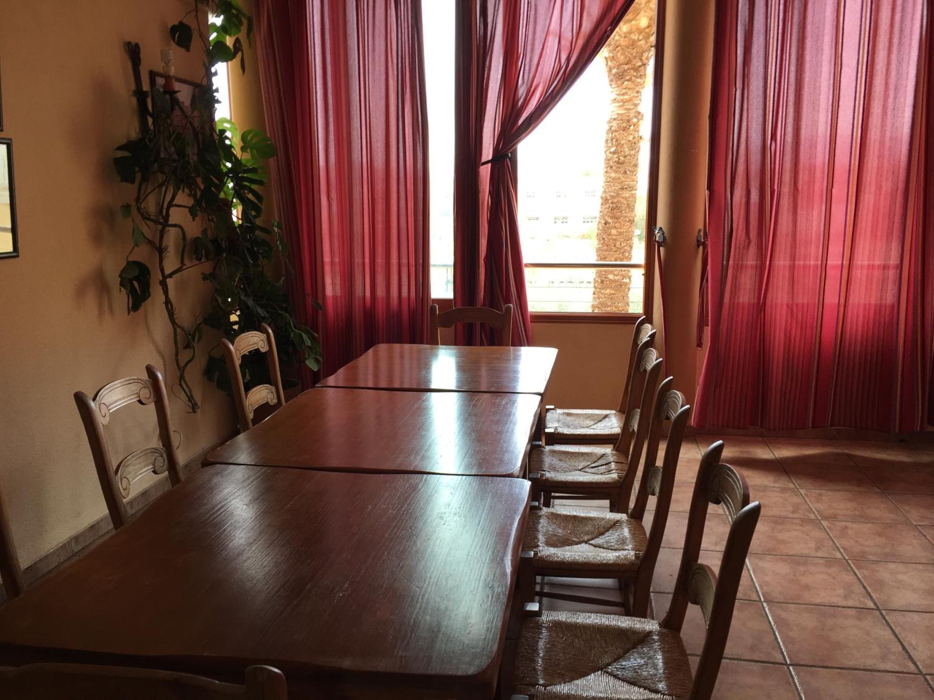 A dining area with a long wooden table and six chairs, red curtains, and a potted plant on the wall.