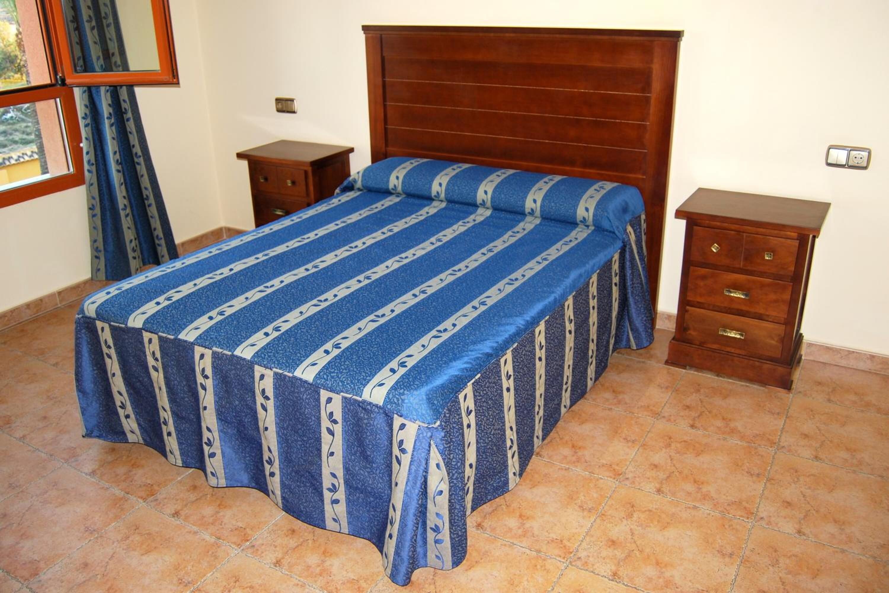 A bedroom with a bed, wooden headboard, two nightstands, a dresser, and a window with curtains.