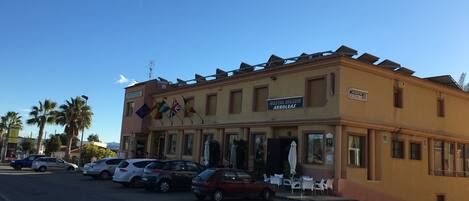 A hostel with a sign reading "HOSTAL MESON ARBOLEAS" and multiple flags. There are cars parked in front and outdoor seating.