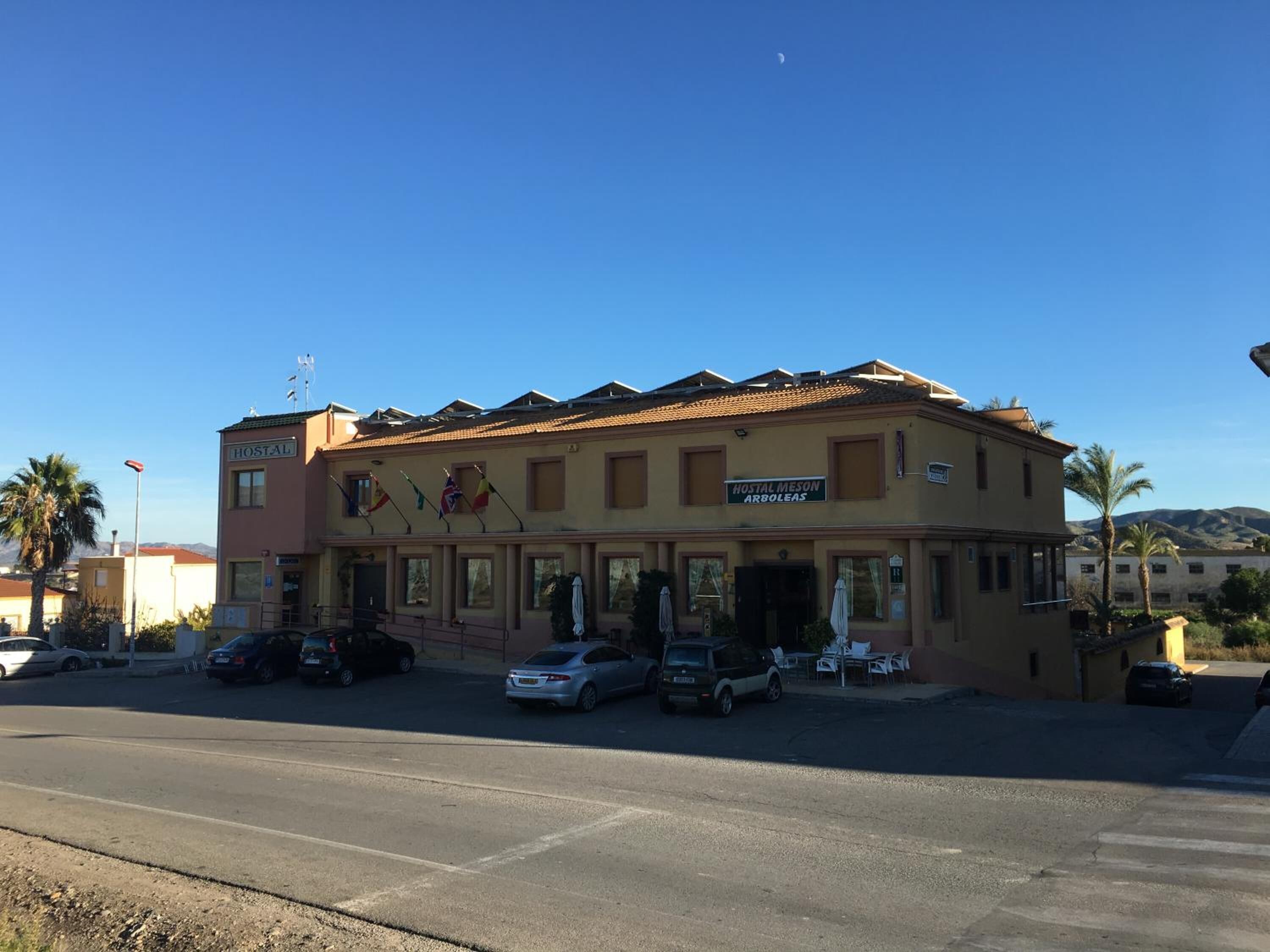 A two-story building with a sign reading "HOSTAL MESON ARBOLEAS" and multiple flags. There are parked cars in front and a clear sky.
