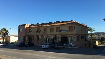 A two-story building with a sign reading "HOSTAL MESON ARBOLEAS" and multiple flags. There are parked cars in front and a clear sky.