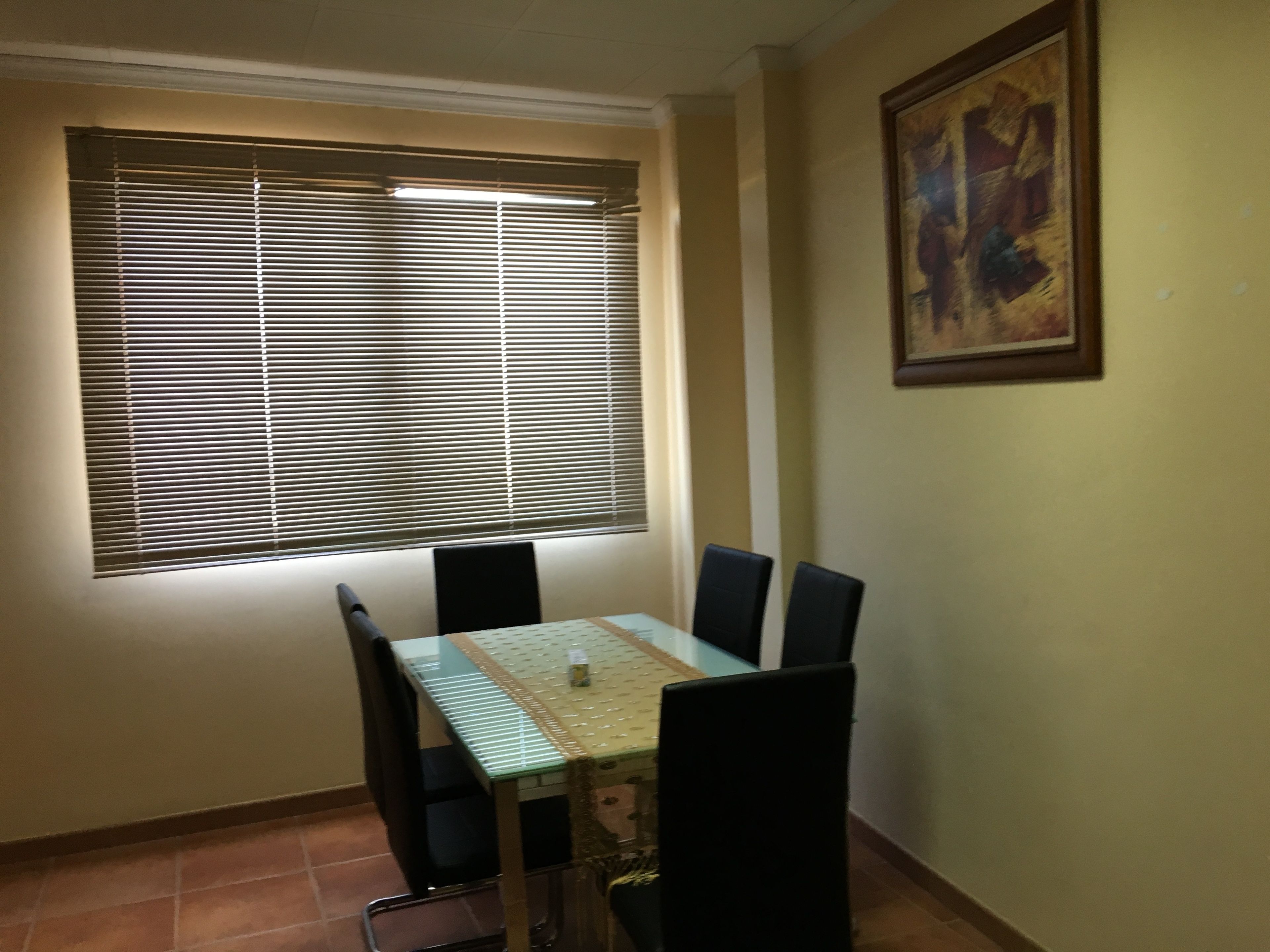 A dining area with a table and chairs, a window with blinds, and a painting on the wall.