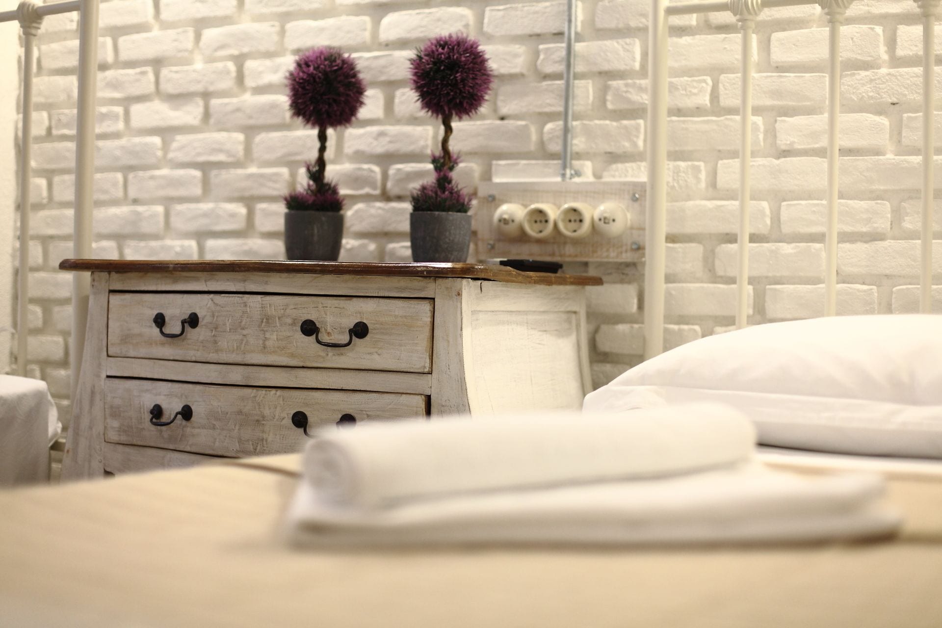Een slaapkamer met een witte bakstenen muur, een houten nachtkastje met twee lades, een bed met witte lakens en twee paarse bloemen op het nachtkastje.